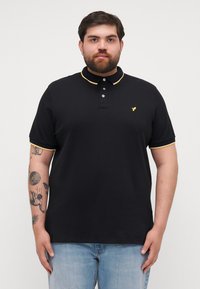 Čierna polo košeľa s žltými akcentmi na golieri a rukávoch, s trojrozmerným zapínaním na gombíky a vyšitým logom na hrudi.