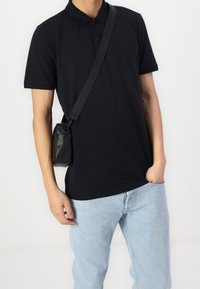 Man med svart polotröja och ljusblå jeans, bärande på en svart crossbody-väska, med handen i fickan, stående mot vit bakgrund.