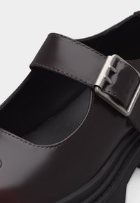 Dunkelbrauner, glänzender Mary-Jane-Schuh aus Leder mit silberner Schnallenriemen und dicker schwarzer Sohle.