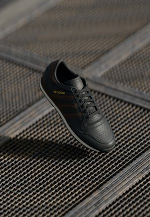 Baskets noires Adidas avec logo jaune et trois bandes sombres, posées en angle sur une surface métallique texturée.