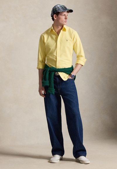 Mladý muž stojí, má na sobě žlutou košili na knoflíky, tmavé džíny, bílé tenisky, zelený svetr uvázaný kolem pasu a károvanou baseballovou čepici.