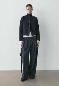 Frau trägt eine schwarze, gerippte Strickjacke mit Knöpfen, weißes Oberteil, lockere dunkle Hose, hält eine schwarze Handtasche, steht vor einer schlichten grauen Wand.