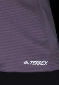 Top deportivo púrpura con tejido texturizado y rayas horizontales, con el logo "TERREX" en blanco y el emblema de Adidas en el dobladillo inferior.