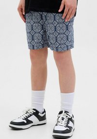 Pantaloni corti blu con un motivo ripetuto intricato, abbinati a scarpe da ginnastica nere e bianche e calzini bianchi alla caviglia.