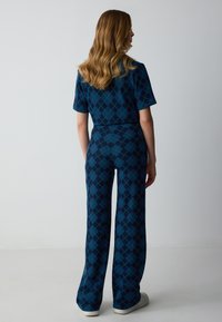 Jumpsuit argyle blu e verde con maniche corte, pantaloni larghi, tessuto testurizzato e vita aderente. La vista posteriore mostra la lunghezza e i dettagli del motivo.