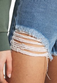 Denim shorts in light blue with frayed hem, featuring multiple distressed cutouts on the thigh. Accompanied by a green sleeve.