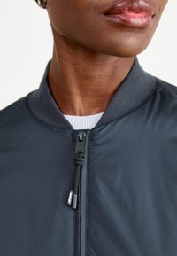 Giacca bomber di un blu navy scuro con collo diritto, chiusura frontale con zip e tessuto texturizzato. Presenta un colletto interno bianco e dettagli con lacci.