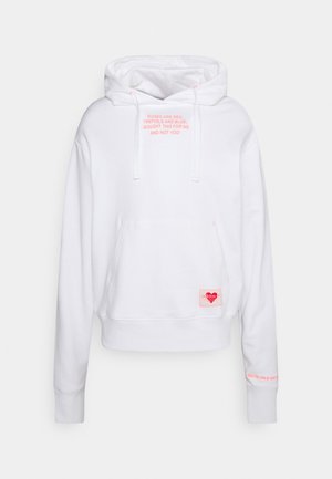 Sweat à capuche blanc avec poche avant, texte rose sur la poitrine affichant un message ludique, et un petit logo cœur rose Adidas près de la poche.