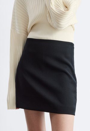 Femme portant une mini-jupe noire et un haut à manches longues côtelé crème avec des poignets évasés, raccourci au niveau de la moitié de la cuisse.