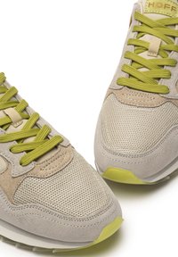 Zapatillas beiges que combinan materiales de malla y ante, con cordones verdes y un diseño de puntera redonda. Los detalles incluyen acentos de color verde claro en la suela.