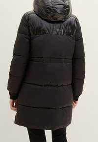 Osoba ubrana w długi czarny pikowany płaszcz z błyszcząca kapturem i górną częścią pleców, stojąca tyłem na tle jasnym.