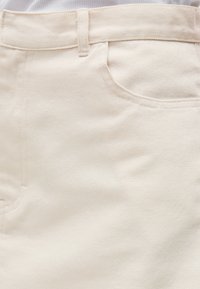 Gros plan d'un pantalon en denim beige montrant la ceinture, la boucle de ceinture et les détails de couture de la poche avant.