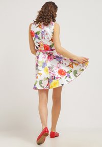 Robe à imprimé floral avec un fond blanc, sans manches, jupe évasée et taille cintrée, accompagnée de chaussures plates rouges.