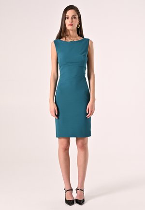 Türkisses knielanges Kleid mit figurbetonendem Schnitt, rundem Ausschnitt und Abnäher. Getragen mit schwarzen spitzen Absatzschuhen und einer goldenen Halskette.
