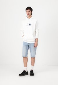 Sweat-shirt blanc à capuche avec un logo, associé à un short en denim bleu clair. Baskets noires, chaussettes blanches, appuyé contre un fond uni.