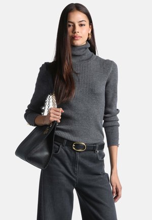 Femme portant un pull à col roulé côtelé gris et un jean noir, tenant un sac à main en cuir noir avec une bandoulière chaîne dorée sur l'épaule.