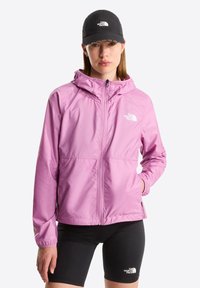 Nainen, joka pukeutunut vaaleanliilaan vetoketjulliseen huppariin, mustaan lippalakkiin ja mustiin shortseihin, kaikki The North Face -logoilla varustettuja.