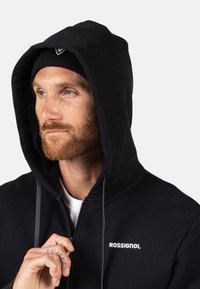 Schwarze Zip-Hoodie mit großer Kapuze, ausgestattet mit einem weißen Logo auf der Brust und Kordeln. Getragen mit einer schwarzen Mütze.
