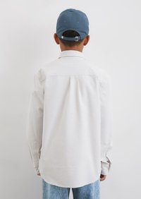 Camicia a maniche lunghe, a righe bianche con orlo curvo, abbinata a un cappello azzurro chiaro, vista da dietro. Il tessuto appare morbido e con texture.