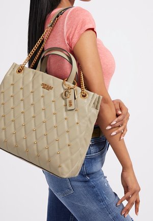Mujer con camisa rosa y jeans azules llevando un bolso beige acolchado de Guess con cadenas doradas y detalles de tachuelas en el hombro.