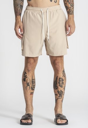 Mand iført beige cargoshorts med hvide snore, sorte slide-sandaler og synlige tatoveringer på ben og arme, stående mod hvid baggrund.