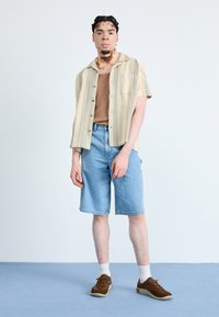 Kurzärmliges, gestreiftes beiges Hemd über einem braunen Tanktop; hellblaue Jeansshorts; braune Turnschuhe mit weißen Akzenten; stehend auf einem blauen Boden.