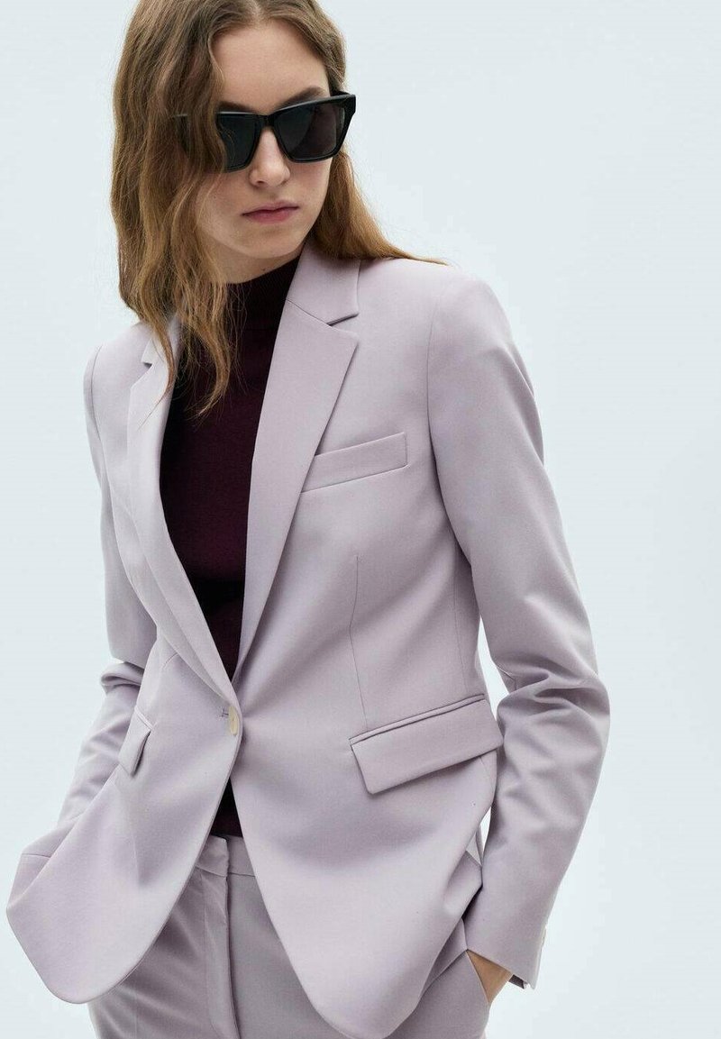 Heller lavendelfarbener Blazer mit taillierter Passform, Reverskragen, seitlichen Taschen und Knopfverschluss. Getragen über einem dunklen Rollkragenpullover, kombiniert mit dunklen Sonnenbrillen.