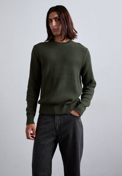 Dark green knit sweater with a crew neckline and ribbed cuffs, paired with grey jeans. The texture appears soft and slightly coarse.