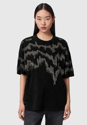 Ung kvinde med langt mørkt hår har en sort oversized T-shirt med sølvperle-lignende abstrakt mønster over bryst og ærmer.