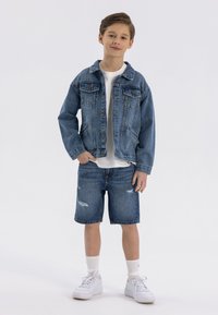 Ensemble en denim bleu clair composé d'une veste et d'un short. La veste comporte une fermeture à boutons et deux poches avant ; le short présente des détails déchirés. T-shirt blanc et baskets.