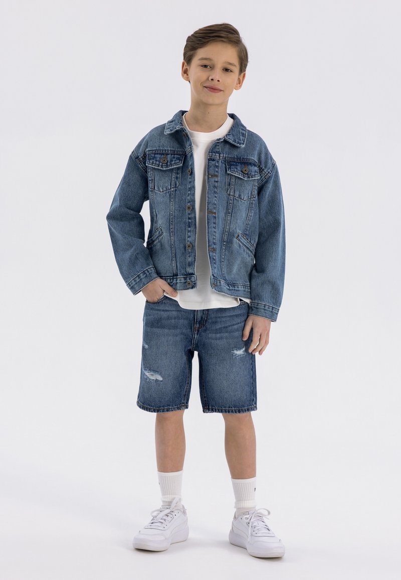 Ensemble en denim bleu clair composé d'une veste et d'un short. La veste comporte une fermeture à boutons et deux poches avant ; le short présente des détails déchirés. T-shirt blanc et baskets.
