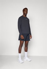 Sportoutfit, bestehend aus einem langärmligen, dunkelgrauen Oberteil und schwarzen Shorts, kombiniert mit weißen Sneakers und Socken. Einfaches Design, ohne Muster.