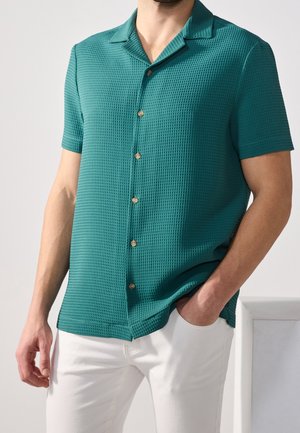 Mężczyzna noszący teksturowaną miętową koszulę z krótkim rękawem i kołnierzykiem, w połączeniu z białymi spodniami, stoi z jedną ręką w kieszeni.