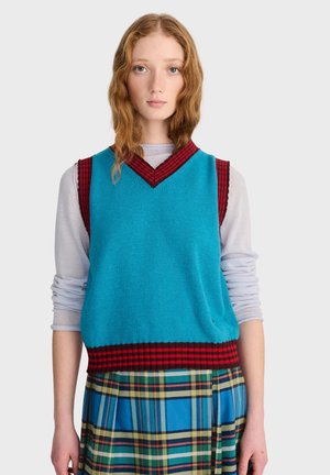 Jeune femme aux cheveux roux ondulés porte un gilet bleu sans manches à rayures rouges par-dessus un haut gris clair à manches longues et une jupe à carreaux bleu-vert.