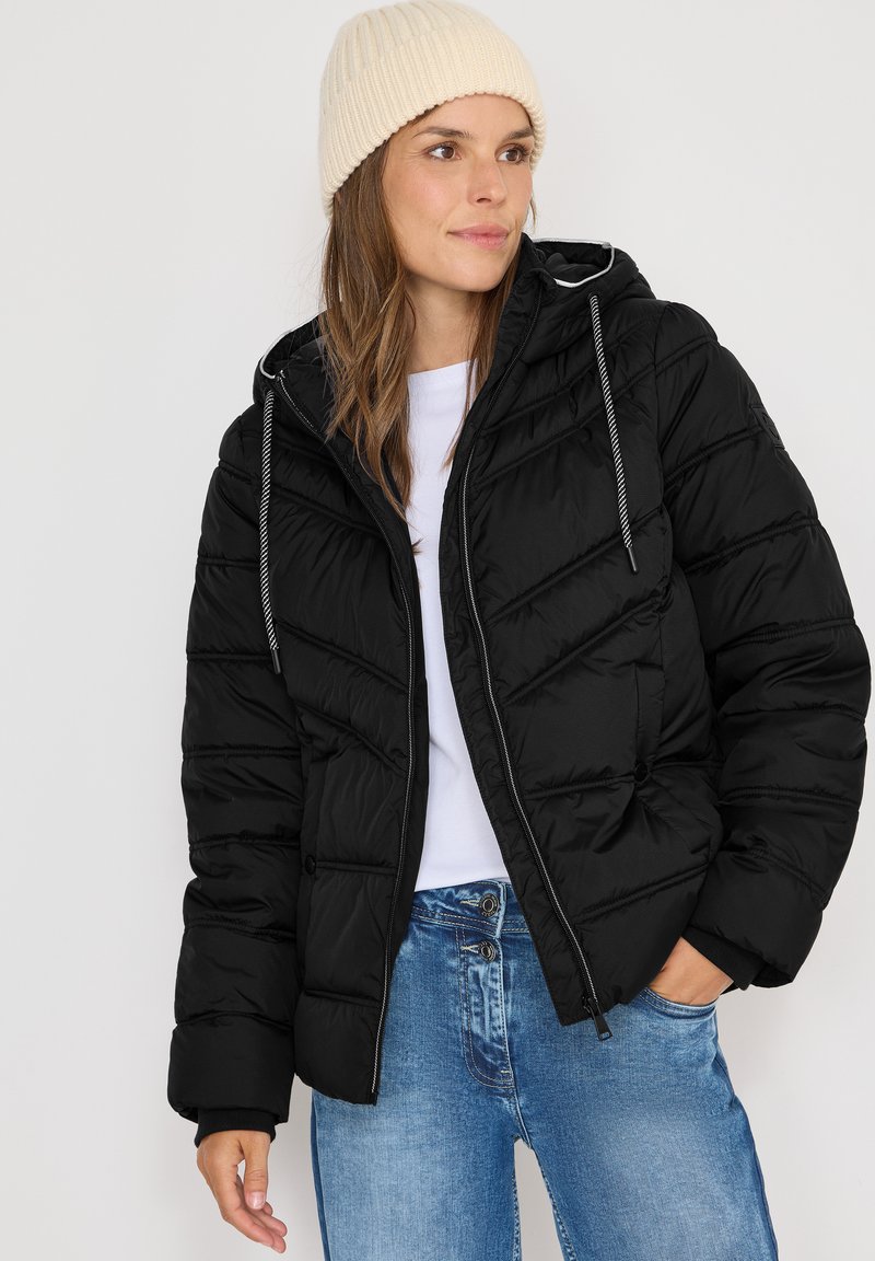Sort pufferjakke med quiltet mønster, lynlåslukning og hætte. Sammen med en hvid skjorte og blå jeans. Lyst farvet strikket hue.