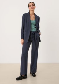 Marineblauwe op maat gemaakte colbert en wijde broek gecombineerd met een getextureerd mintgroen top. Glanzende zwarte schoenen maken de look compleet.