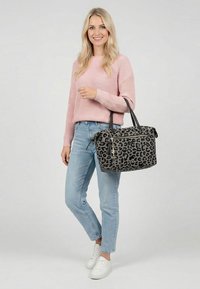 Frau in pinkem Pullover und hellblauen Jeans, die eine große graue Leopardenmuster-Handtasche mit schwarzen Griffen hält und vor einem schlichten Hintergrund lächelt.