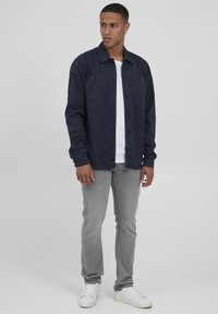 Camicia button-up blu navy con due tasche sul petto, abbinata a jeans grigi e sneaker bianche. Il tessuto sembra liscio e leggero.