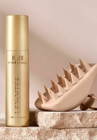 Gul sprayflaska märkt "Håraktiverande serum" bredvid en beige massager med spetsig yta, placerad på en strukturerad stenbakgrund.