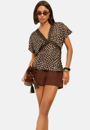 Kvinde iført solbriller, en brun prikket bluse med blonder, brune shorts, holder en brun taske med kvaster og bærer armbånd på det ene håndled.