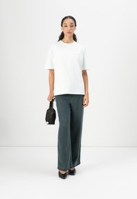 White cotton t-shirt with short sleeves, paired with dark green wide-leg jeans. Model holds a small black bag, standing against a neutral background.