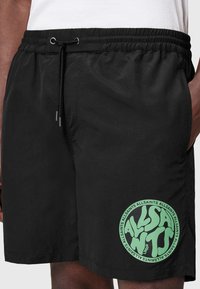 Bañadores negros con cinturilla elástica y cordón, que presentan un logo circular verde con texto estilizado en la pierna izquierda.