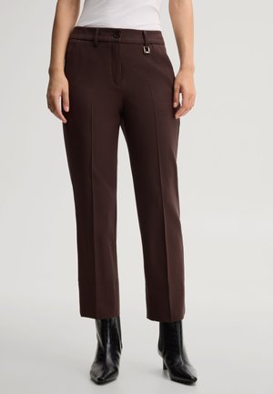 Donna che indossa pantaloni marrone scuro su misura alla caviglia con pieghe frontali e stivaletti neri con tacco alto, in piedi davanti a uno sfondo semplice.