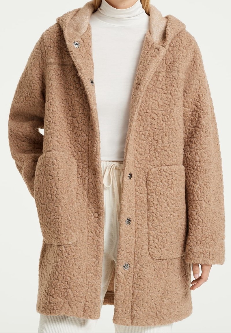 Manteau en laine beige texturé avec grandes poches avant, boutons-pression, porté sur un col roulé blanc et un pantalon clair.