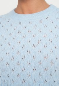 Ανοιχτό μπλε πλεκτό πουλόβερ με σχέδιο κυμάτων, με ριμπ ζώνη στο λαιμό και ανοιχτό πλεκτό ύφασμα. Κοντινή λήψη των λεπτομερειών του υφάσματος.