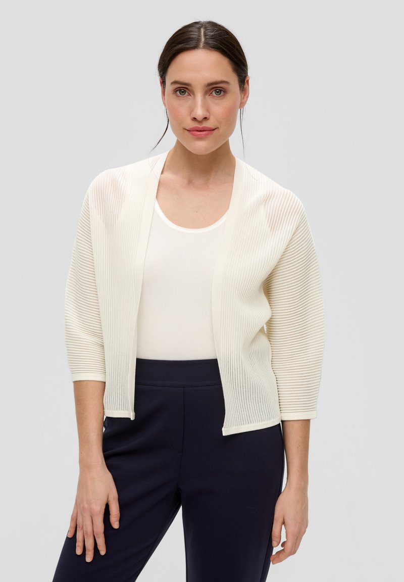 Lichte, geribde cardigan in off-white, open voorkant met driekwartmouwen, gecombineerd met een aansluitend wit topje en donkerblauwe broeken.
