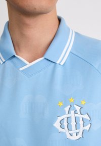 Helehelesin sinine polo särk pehme tekstuuriga, millel on valge triibuline krae, bordüüritud logo ja kolm kollast tähte disaini kohal.