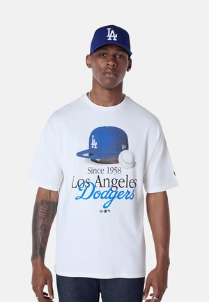 Hombre que lleva una camiseta blanca de Los Angeles Dodgers con gorra azul y gráfico de béisbol, y una gorra azul de LA Dodgers puesta hacia adelante.
