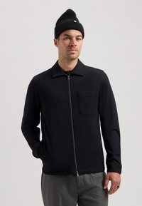 Chaqueta negra con cremallera, cuello, mangas largas y un bolsillo en el pecho. Textura suave, combinada con pantalones grises y un gorro de punto negro.