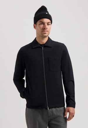 Black zip-up jacket with a collar, long sleeves, and a chest pocket. Smooth texture, paired with gray trousers and a black knit beanie.